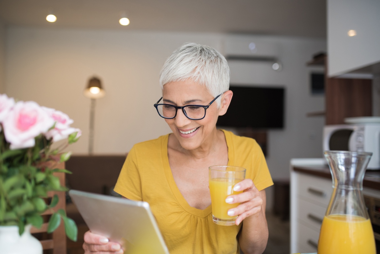 Woman in yellow, drinking orange juice looking at her tablet and smiling