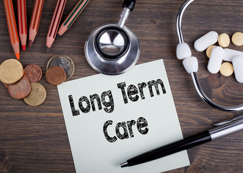 Note reading “Long Term Care” on a desk surrounded by coins, pencils, a stethoscope, and medication, representing healthcare and financial planning costs