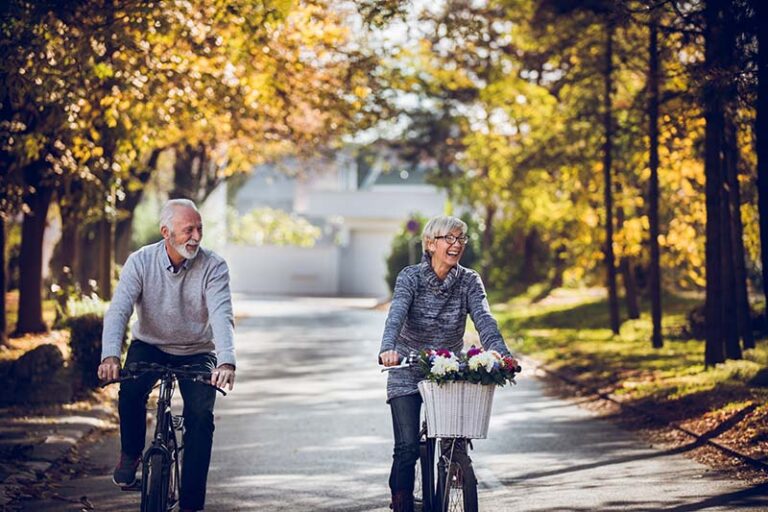 A retired couple riding bicycles down a quiet, tree-lined street in the fall. 