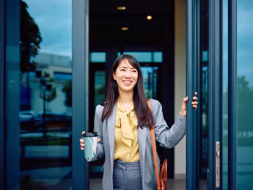 A businesswoman walks out of an office building with a coffee in hand.